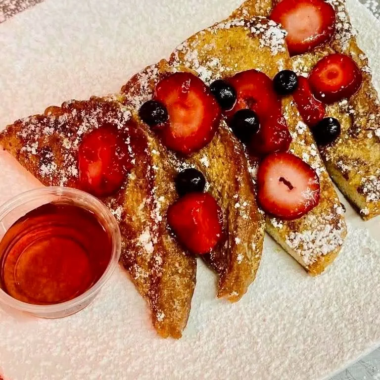 Powdered-sugar French toast topped with strawberries and blueberries, with a small cup of syrup